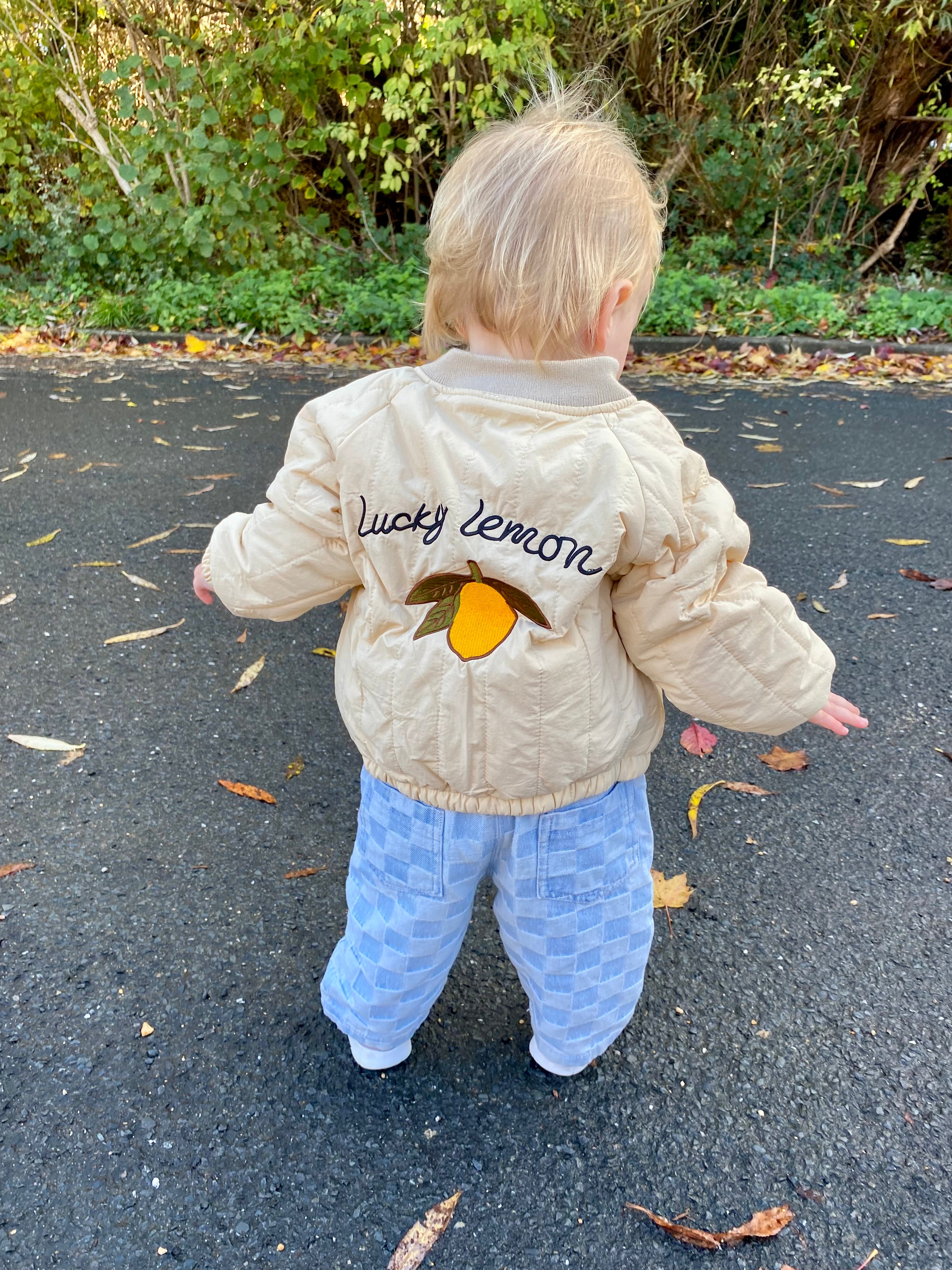 Child wearing a beige jacket with 'Lucky Lemon' branding outdoors on a leafy path.